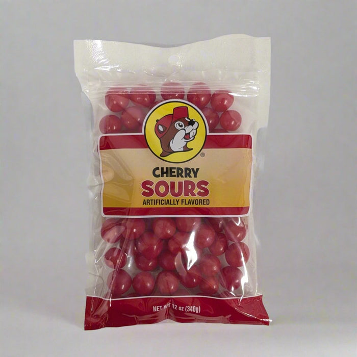 A clear plastic bag of "Cherry Sours" candy is displayed on a white surface. The bag features a red and yellow label with a cartoon character resembling a beaver wearing a hat. The candies inside are small, round, and bright red, indicating the cherry flavor. The packaging specifies a net weight of 12 ounces (340 grams).